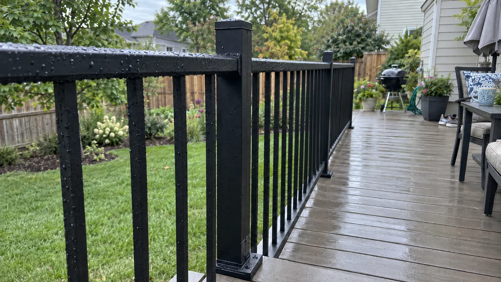 A dark metal railing after rainfall, with square spindles evenly set and the post foot plate fixed onto the boards